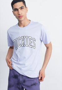 Svetle levandulové bavlněné tričko s kulatým výstřihem a výrazným černým logem "DICKIES" na přední straně. Nošeno s fialovými kalhotami s tie-dye vzorem.