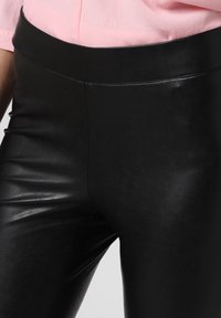 Svarta leggings i faux läder med hög midja, slät textur och inga synliga sömmar, kombinerade med en ljusrosa skjorta vid midjan.