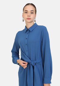 Blue button-up dress with long sleeves and a tie waist detail. Smooth fabric, collar, and knotted accent at the waist.