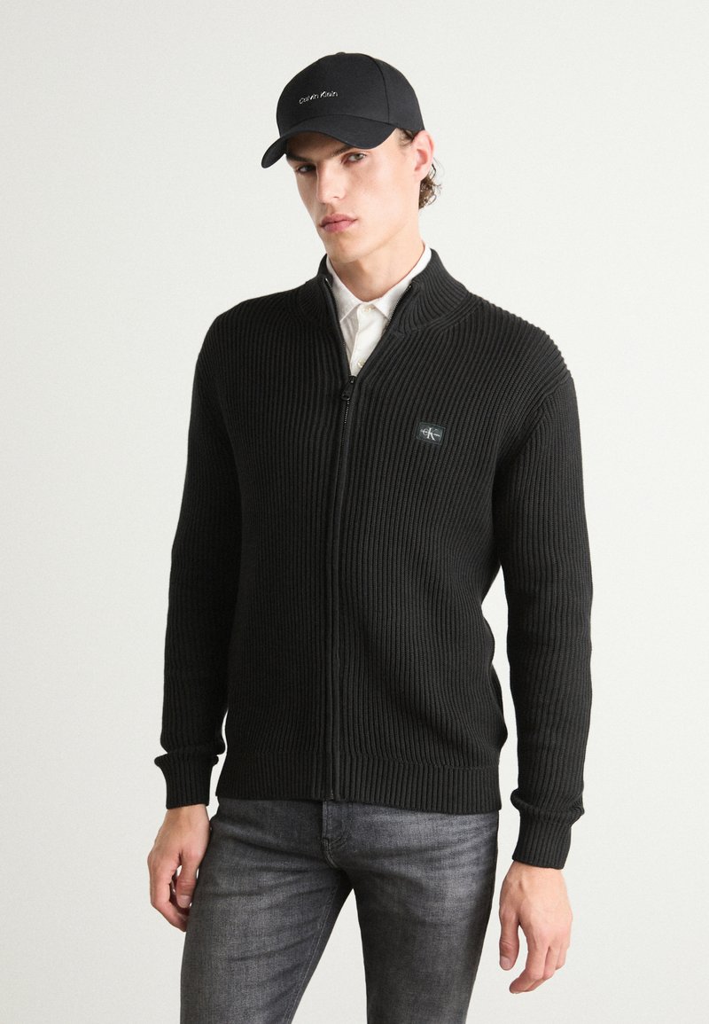 Maglione a zip nero a coste con colletto alto, dotato di una discreta toppa con logo. Indossato sopra una camicia bianca, abbinato a jeans scuri e un cappellino nero.