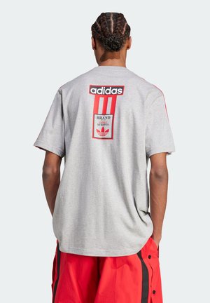 T-shirt en coton gris avec trois bandes rouges et logo au dos. Coupe ample avec manches courtes et accents rouges contrastants sur les épaules.