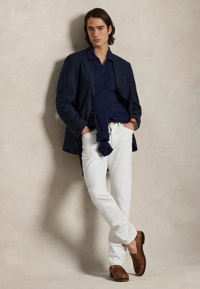 Νέος άνδρας που ακουμπάει σε τοίχο φορόντας μπλε ναυτικό σακάκι, μπλε πουκάμισο, λευκό παντελόνι, καφέ δερμάτινα παπούτσια και ρολόι, με τα χέρια στις τσέπες.