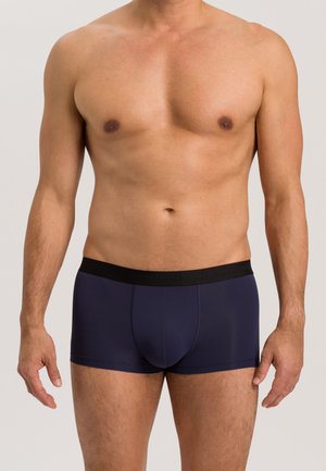 Torace maschile con boxer blu navy con fascia nera in vita su sfondo chiaro uniforme, braccia rilassate ai lati.