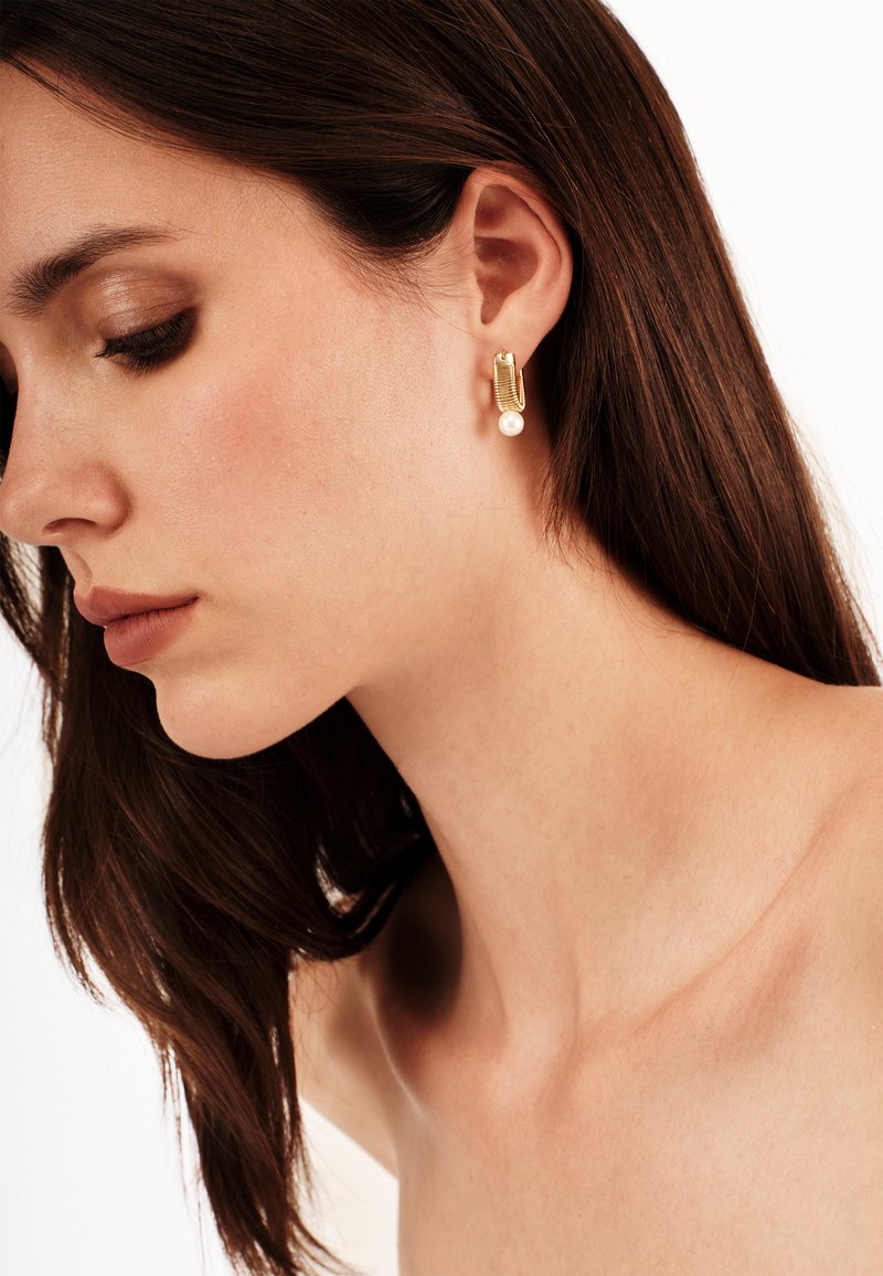 Orecchini in tono oro con un design a cerchio texturizzato e un accento di perla bianca staccata. Indossati sull'orecchio sinistro di una donna con i capelli lunghi.