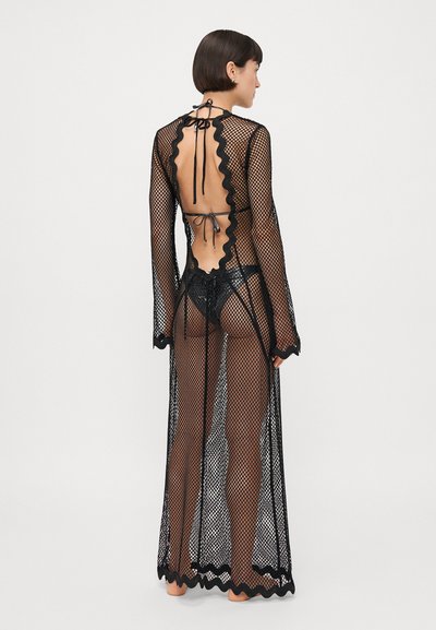 Robe longue en filet noir avec des bords festonnés. Design à dos ouvert, manches longues et fermeture à nouer au cou. Texture translucide tout au long.