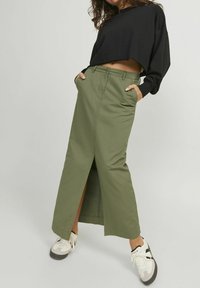 Falda larga verde oliva con una abertura frontal, que presenta bolsillos laterales y una cinturilla ajustada, combinada con un suéter corto negro y zapatillas.
