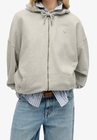 Felpa grigia in cotone con zip, cappuccio regolabile, dotata di cerniera frontale, tasche laterali e un piccolo logo sul petto.