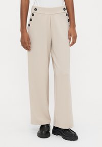 Μπεζ παντελόνια με φαρδιά μπατζάκια και μαύρα κουμπιά στο πλάι, φορεμένα με μαύρες chunky μοκασίνια, σε ένα άτομο που στέκεται μπροστά σε απλό φόντο.