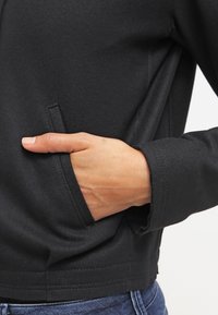 Main partiellement dans la poche latérale d'une veste noire portée sur un jean bleu.