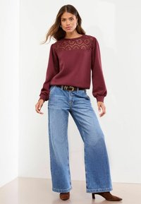 Bordo dukserica z detajli čipke na ramenih, uparena sa širokim plavim farmerkama i smešnim čizmama na petu. Glatka tekstura, ležeran dizajn.