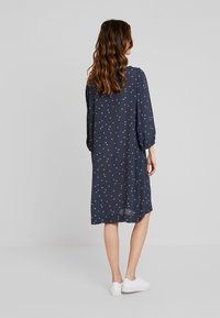 Námořnické modré šaty s bílými puntíky, dlouhými rukávy a volným střihem, ve spojení s bílými teniskami, ukazujícími lehkou texturu látky.