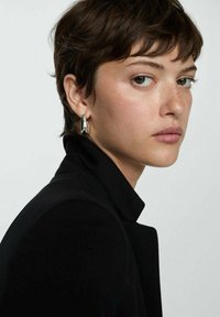 Close-up of a person with short brown hair, wearing a black blazer. Notable silver hoop earring, natural skin texture, and light makeup.