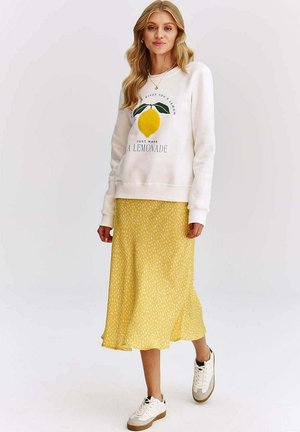 Biela mikina so grafickým motívom citróna a textom, v kombinácii so žltou sukňou midi s bodkami. Celkom outfit dopĺňajú biele tenisky s hnedými podrážkami.