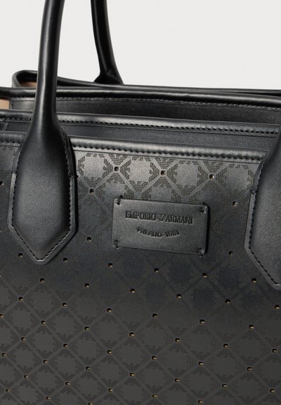 Μαύρη δερμάτινη τσάντα Emporio Armani με διακοσμητικές τρύπες, δύο χερούλια και μαύρη ετικέτα με την επωνυμία "Emporio Armani Milano 1981".