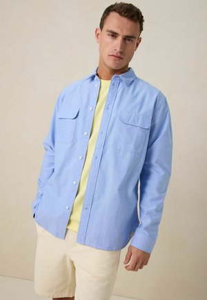 Lichtblauw button-up overhemd met twee borstzakken, gedragen over een lichtgele t-shirt, gecombineerd met crèmekleurige shorts. Gladde katoenen stof.