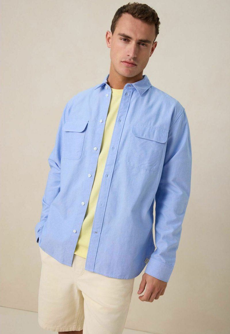 Lichtblauw button-up overhemd met twee borstzakken, gedragen over een lichtgele t-shirt, gecombineerd met crèmekleurige shorts. Gladde katoenen stof.