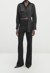 Person trägt eng anliegende schwarze hohe Hosen, ein kurzes schwarzes Hemd mit Knöpfen und einem langen dekorativen Schal sowie spitz zulaufende schwarze Schuhe.