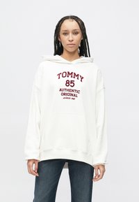 Biela mikina s kapucňou s veľkým bordovým logom "TOMMY" a textom, voľný strih a predné vrecko. Nosená s modrými džínsami.
