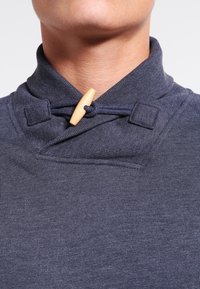 Tmavě modrý bavlněný top s falešným rolákem, vybavený dřevěným zapínáním na kličku a jemným texturovaným vzorem, se šitými detaily na límci.