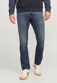 Jeans de mezclilla azul oscuro con corte recto, que presentan áreas descoloridas y costuras en contraste, combinados con zapatillas blancas y un sudadera azul marino.