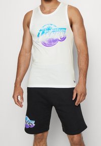 Biała koszulka na ramiączkach z grafiką "Los Angeles Lakers" w niebieskim i fioletowym, zestawiona z czarnymi szortami z logo na udzie. 