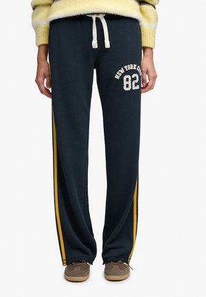 Personne portant un pantalon de survêtement bleu marine avec des bandes latérales jaunes, un cordon blanc, le texte "New York City 82", des chaussures marron et un pull jaune.