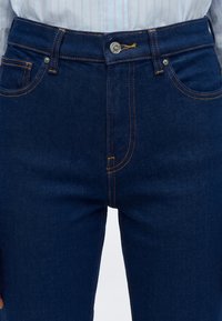Gros plan d'une personne portant un jean en denim bleu foncé avec des coutures orange et un bouton argenté, associé à une chemise claire à rayures.