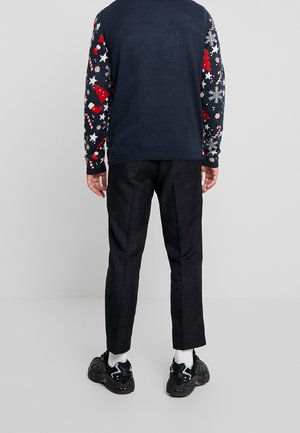 Pull marine avec des motifs festifs en rouge, blanc et bleu ; pantalon noir texturé ; baskets noires avec des accents blancs.