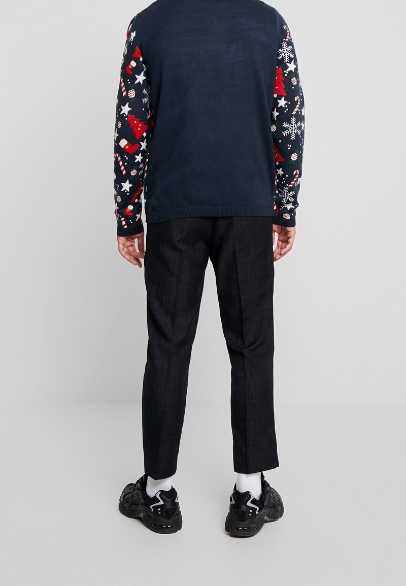 Pull marine avec des motifs festifs en rouge, blanc et bleu ; pantalon noir texturé ; baskets noires avec des accents blancs.