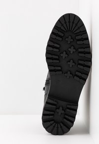 Semelle de botte noire avec un motif de crampons profond et robuste comprenant des crampons épais et des indentations en forme de croix sur fond blanc.
