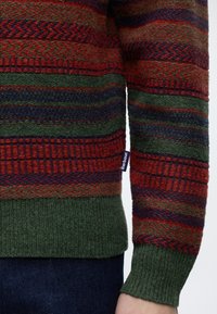 Gros plan sur la manche et l'ourlet d'un pull en laine à motifs verts, rouges et bleu foncé avec une petite étiquette de la marque Barbour, porté sur un jean en denim foncé.