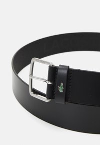 Black leather belt with a silver metal buckle. Features a Lacoste logo and a green alligator accent on the strap. Smooth texture and minimalist design.