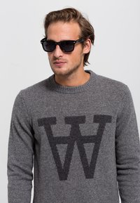 Pulover tricotat gri cu un logo mare de culoare gri închis "W" pe partea din față, purtat cu ochelari de soare negri, evidențiind designul de modă casual.