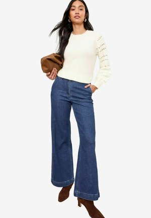 Cremfarbener Pullover mit Lochmuster-Details an den Ärmeln, kombiniert mit weit geschnittenen blauen Jeans. Hält eine braune Tasche und trägt braune Stiefeletten mit Absatz.