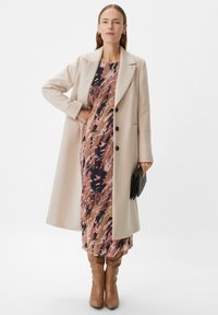 Beige Wollmischungs-Mantel mit Reverskragen und drei Knöpfen, über einem rosa, braunen und marineblau gemusterten Kleid getragen. Braune kniehohe Stiefel.