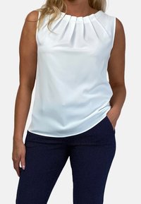 Kvinna som bär en ärmlös vit plisserad blus och marinblå byxor med små vita prickar, stående med en hand i fickan.