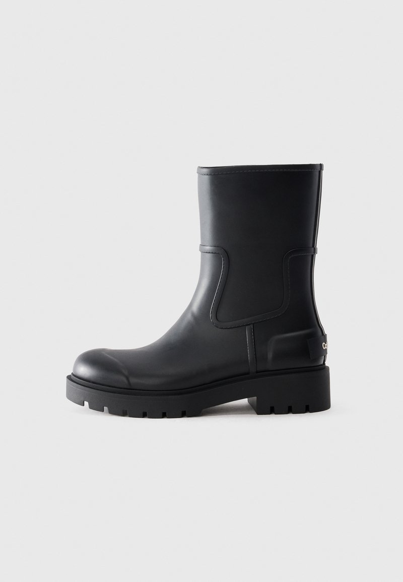 Botte en caoutchouc noire mi-mollet avec semelle à crampons épaisse, bout rond et languette à l'arrière sur fond clair.