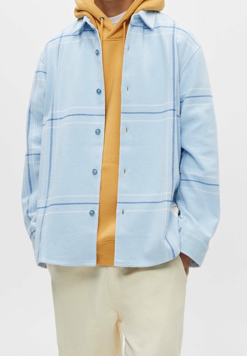 Camicia a quadri blu chiaro con accenti blu, con bottoni davanti, indossata sopra un hoodie senape. Tessuto morbido, vestibilità rilassata, maniche corte.