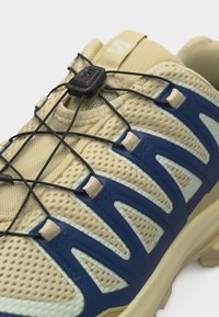 Μπεζ αθλητικό παπούτσι από δίχτυ με επενδύσεις σε ναυτικό μπλε, μαύρο ελαστικό σύστημα γρήγορης δέσης και θηλιά στο γλώσσα.