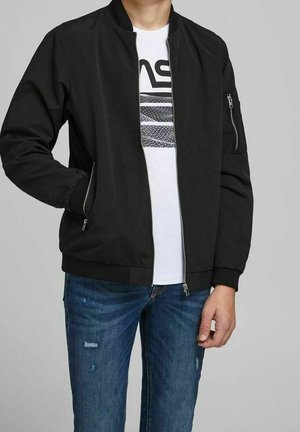 Person iført en sort bomberjakke med lynlås over en hvid T-shirt med grafik og blå slidte jeans, stående foran en ensfarvet grå baggrund.