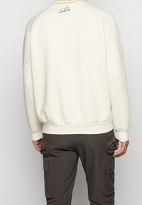 Cream-colored sweatshirt with ribbed cuffs and hem, featuring a signature detail on the back. Made from soft, textured fabric.