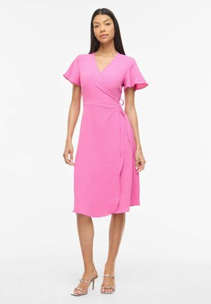 Kvinne iført en knelengde rosa omslagskjole med korte flagreermer og sølvfargede høye hæler, stående mot en hvit bakgrunn.