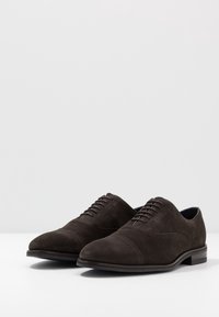 Chaussures habillées en daim marron foncé avec une texture lisse, un bout pointu et six œillets pour les lacets. Talon plat et détails de couture minimaux.