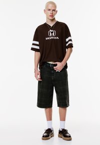 Bruine oversized t-shirt met witte Honda-logo en strepen, gecombineerd met donkere geruite shorts. Draagt bruin schoeisel en witte sokken.