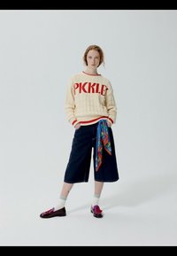 Jeune personne debout, portant un pull crème avec le texte "PICKLE", un pantalon culotte bleu marine, une ceinture écharpe colorée, des chaussettes blanches et des chaussures bordeaux.