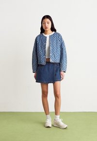 Sinine disainiga denim-jakk, millel on vatiiniga tekstuur, kantud valge särgi ja tumeda denim-seeliku peal. Modell kannab valgeid sneakers'e ja pahkluuni sokke.