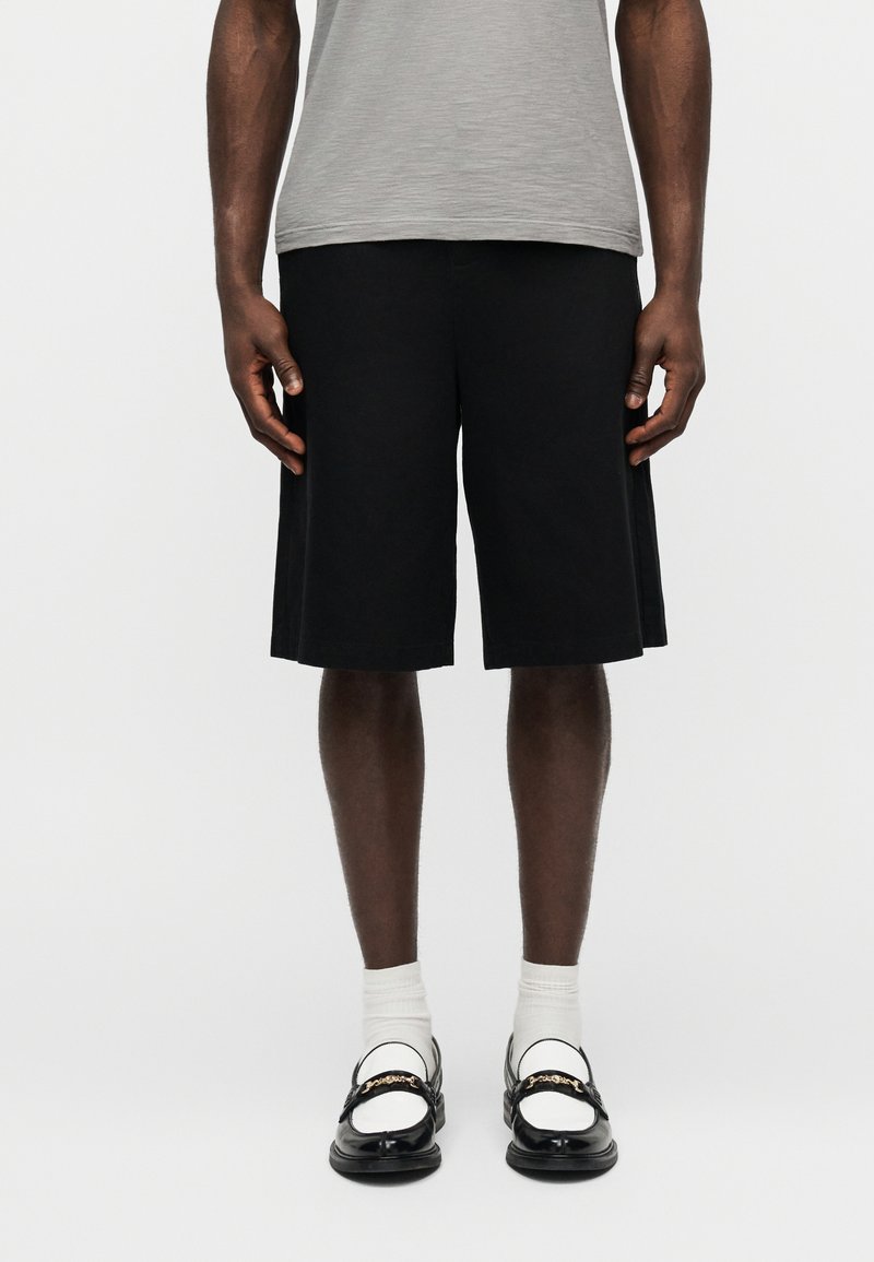 Άνδρας που φοράει μαύρα σόρτς μέχρι το γόνατο, λευκές κάλτσες, μαύρα και λευκά μοκασίνια και ανοιχτό γκρι μπλουζάκι, στέκεται μπροστά σε φόντο απλό.