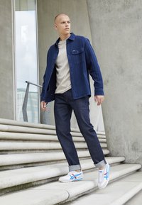 Neformální outfit sestávající z tmavě modré košile s kapsami a světle šedého trička, doplněný tmavými džínami a bílými teniskami s modrými akcenty.