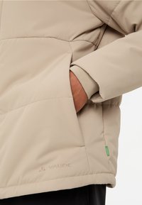 Giacca imbottita beige con tasca zip, polsini elasticizzati e etichetta verde. Presenta cuciture sottili e un logo vicino all'orlo.