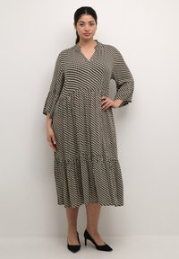 Schwarz-weißer, gemusterter Kleid mit einem gestuften Saum, langen Ärmeln und einem kragenlosen Ausschnitt, aus leichtem Stoff. Kombiniert mit schwarzen Absatzschuhen.